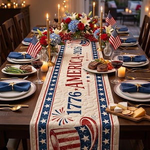 May include: A festive dining table set for a patriotic celebration. The table features a cream-colored runner with red and blue stars and stripes, and the text "1776 AMERICA 2026".  Plates, napkins, and floral arrangements complete the scene.