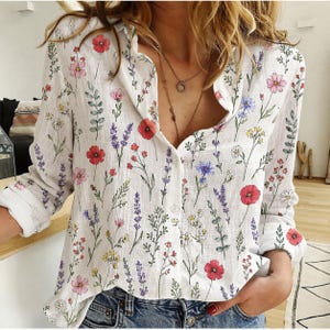 May include: White button-down shirt with a colorful floral print. The shirt features a variety of flowers in red, pink, purple, and blue, with green leaves. The long-sleeved shirt is made of a lightweight fabric.