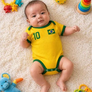 Puede incluir: Un body de bebé amarillo con bordes verdes, que presenta el número 10 y una bandera de Brasil en el pecho. El body está rodeado de varios juguetes coloridos, incluyendo una pelota amarilla, un juego de anillos apilables y figuras de peluche.