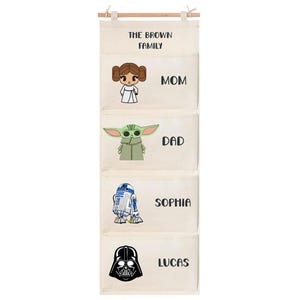 May include: A beige fabric wall hanging organizer with four pockets, featuring the text THE BROWN FAMILY at the top. Each pocket displays a character illustration and a name: MOM with a princess, DAD with a green alien, SOPHIA with a robot, and LUCAS with a black helmeted figure.