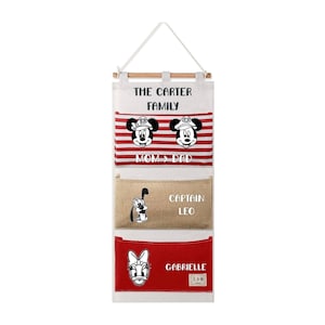 May include: A hanging wall organizer with three pockets. The top pocket has red and white stripes with cartoon mouse characters and the text THE CARTER FAMILY MOM & DAD. The middle pocket is tan with a cartoon dog and CAPTAIN LEO. The bottom is red with a cartoon duck and GABRIELLE.