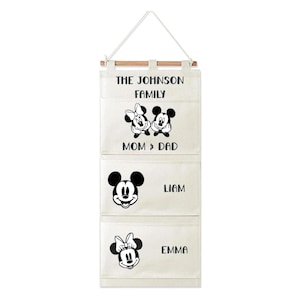 May include: A hanging canvas wall organizer with three pockets, suspended by a wooden rod and white cord. The fabric features black printed text reading THE JOHNSON FAMILY, MOM & DAD, LIAM, and EMMA, alongside black graphic illustrations of Mickey and Minnie Mouse.