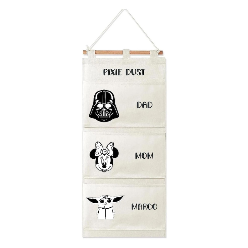 May include: A cream-colored fabric wall organizer with three pockets hanging from a wooden dowel and white cord. The top pocket features the text PIXIE DUST. Below are black graphic prints of Darth Vader labeled DAD, Minnie Mouse labeled MOM, and Grogu labeled MARCO.