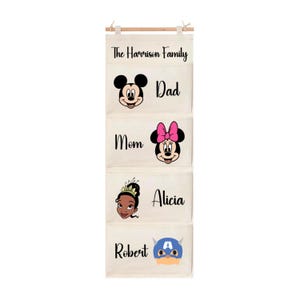 Puede incluir: Organizador colgante de tela beige con cuatro bolsillos. El bolsillo superior dice "The Harrison Family". Debajo hay bolsillos etiquetados como "Dad" con un gráfico de Mickey Mouse, "Mom" con un gráfico de Minnie Mouse, "Alicia" con un gráfico de princesa y "Robert" con un gráfico del Capitán América.