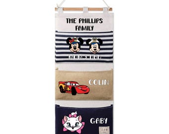 May include: A multi-pocket wall organizer with a wooden dowel and hanging cord. The top pocket has the text "THE PHILLIPS FAMILY" above a striped section with Mickey and Minnie Mouse. Below are pockets with the names "COLIN" and "GABY" and character graphics.