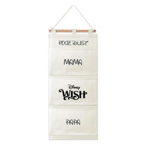 Può includere: Un organizer da parete sospeso in tela color crema con tre tasche, appeso a un bastoncino di legno e a un cordino bianco. Le tasche presentano scritte nere che riportano Pixie Dust, Mama, Disney Wish e Papa in vari caratteri decorativi.