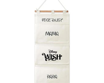 May include: A cream-colored canvas hanging wall organizer with three pockets suspended from a wooden dowel and a white cord. The pockets feature black text reading Pixie Dust, Mama, Disney Wish, and Papa in various decorative fonts.