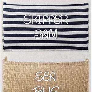 Könnte beinhalten: Ein vertikaler Wand-Organizer mit drei Taschen, der an einem Holzstab mit einem weißen Seil aufgehängt ist. Die obere Tasche ist marineblau-weiß gestreift und mit dem Text SKIPPER SAM versehen. Die mittlere Tasche ist beige, und die untere marineblaue Tasche zeigt den Text LIL SAILOR sowie ein kleines Etikett mit einem Anker-Symbol.