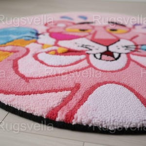Op de afbeelding: Een ronde, roze vloerkleed met een cartoonontwerp van de Roze Panter. Het vloerkleed heeft een zwarte rand en is gemaakt van een zacht, getextureerd materiaal. De Roze Panter wordt afgebeeld met een glimlachende uitdrukking.