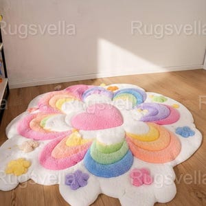 May include: A colorful, cloud-shaped rug with a rainbow and flower design. The rug features a white base with pink, yellow, orange, blue, and purple accents. The rug is made of a soft, plush material and is suitable for a child's room or playroom.