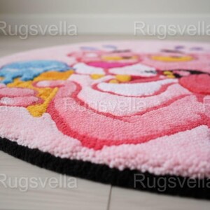 Op de afbeelding: Een ronde, roze vloerkleed met een cartoon-personage ontwerp. Het kleed heeft een zwarte rand en een getextureerd oppervlak. Het ontwerp bevat roze, gele en blauwe tinten. Geschikt voor een kinderkamer.