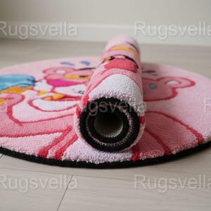 Op de afbeelding: Een ronde roze vloerkleed met het stripfiguur Pink Panther, gedeeltelijk opgerold. Het kleed heeft een zwarte rand en een getextureerd oppervlak. Het ontwerp bevat roze, rode en blauwe tinten.