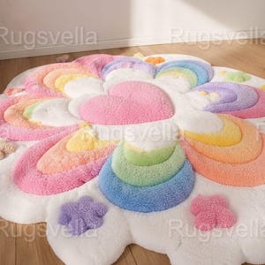 May include: A round, fluffy rug in a whimsical design. The rug features a white base with colorful accents, including a pink heart, rainbow arcs, and cloud-shaped details. The rug is made of soft, plush material.
