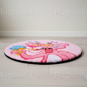 Op de afbeelding: Een ronde roze vloerkleed met het stripfiguur Pink Panther die een ijsje vasthoudt. Het vloerkleed heeft een zwarte rand en ligt op een lichte houten vloer. De Pink Panther is in roze en rode tinten.