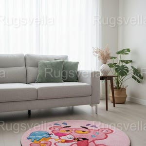 Op de afbeelding: Een ronde roze vloerkleed met een cartoon Pink Panther die een ijsje vasthoudt. Het kleed heeft een zwarte rand en ligt op een lichte houten vloer. De achtergrond toont een grijze bank, groene kussens en een plant in een pot.
