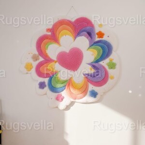May include: A colorful, handmade rug in a flower shape, featuring a pink heart center and rainbow-colored petals. The rug is adorned with small, decorative flower accents in various colors and is mounted on a white wall.