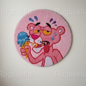 Op de afbeelding: Een roze ronde vloerkleed met een cartoonafbeelding van de Roze Panter die een blauw ijsje vasthoudt. De Roze Panter zweet, draagt een gele bril en steekt zijn tong uit. Een leuk decoratief item.