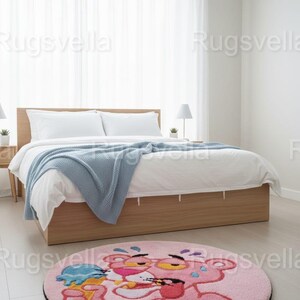 Op de afbeelding: Een ronde roze vloerkleed met het stripfiguur Pink Panther die ijs eet. Het vloerkleed heeft een zwarte rand en ligt op een lichte houten vloer in een slaapkamer. Op de achtergrond staat een bed met wit beddengoed en een blauwe deken.