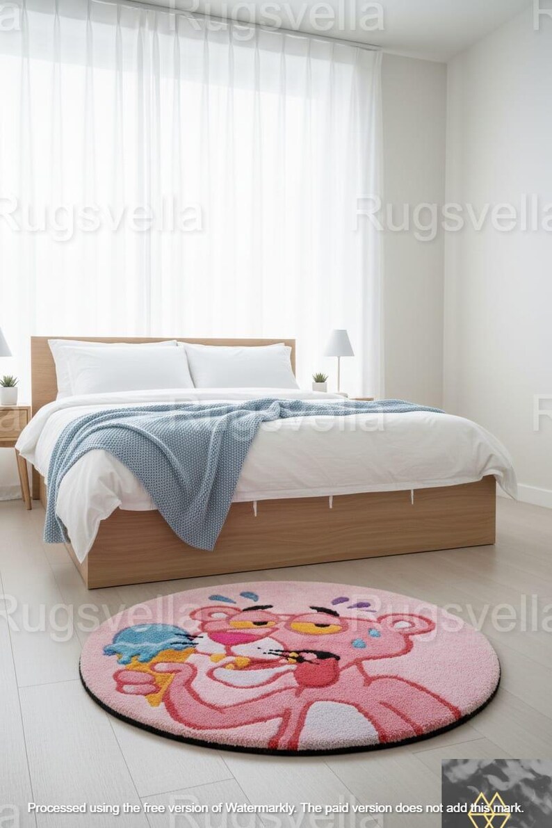 Op de afbeelding: Een ronde roze vloerkleed met het stripfiguur Pink Panther die ijs eet. Het vloerkleed heeft een zwarte rand en ligt op een lichte houten vloer in een slaapkamer. Op de achtergrond staat een bed met wit beddengoed en een blauwe deken.