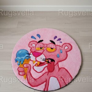Op de afbeelding: Een ronde roze vloerkleed met een cartoon Pink Panther-personage met een bril en een blauw ijsje. Het vloerkleed heeft een zwarte rand en ligt op een lichte houten vloer. De diameter van het vloerkleed is ongeveer 100 cm.