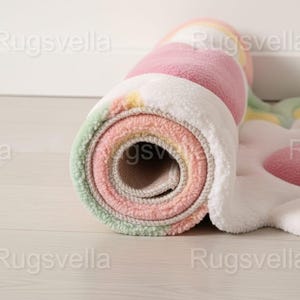 May include: Rolled-up, colorful rug with a flower design. The rug features pastel colors including pink, green, yellow, and white. The rolled section reveals the layers of the rug, showcasing its soft texture and design.