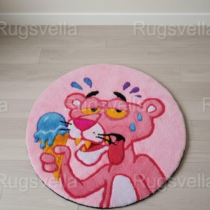 Op de afbeelding: Een ronde roze vloerkleed met een cartoon Pink Panther-personage met een bril en een blauw ijsje. Het vloerkleed heeft een zwarte rand en ligt op een lichte houten vloer. De diameter van het vloerkleed is ongeveer 100 cm.