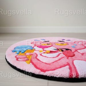 Op de afbeelding: Een ronde roze vloerkleed met de tekenfilmfiguur Pink Panther die een blauw ijsje vasthoudt. Het vloerkleed heeft een zwarte rand en ligt op een lichte houten vloer. Het woord "Rugsvella" staat als watermerk op de afbeelding.