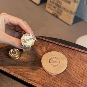May include: A hand holding a gold-colored stamp with the words "SOLLIEBA HANDMADE" around the edge. A second stamp and a stamped leather coaster are also visible on a wooden surface. The coaster has the words "SOLLIEBA POTTERY" stamped on it.