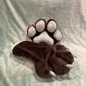 May include: A brown plush paw with light blue pads and claws, and a matching paw-shaped slipper. The items are designed to resemble a bear or other animal paw. The background is a light blue fabric.