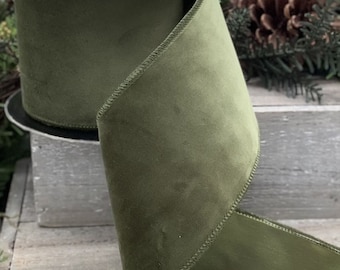 May include: A wide spool of olive green velvet ribbon with a soft, matte texture and finished stitched edges. The ribbon is wound around a central core, with a portion of the fabric cascading downward, displaying a consistent deep green color.