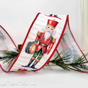 May include: White ribbon with a red border featuring a nutcracker soldier design. The soldier wears a red coat, a tall hat, and holds a drum. The ribbon is decorated with musical notes and is suitable for Christmas decorations.
