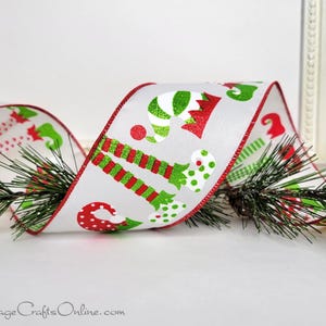 May include: White Christmas ribbon with red trim and festive elf leg and candy cane designs in red and green. The ribbon is wrapped around a sprig of green pine needles, creating a holiday theme.