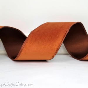May include: A length of orange velvet ribbon with a brown underside, stitched along the edges. The ribbon is curled in a wave pattern, showcasing its texture and color. The ribbon is likely used for crafts, gift wrapping, or decorative purposes.