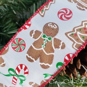 May include: A white fabric ribbon with a repeating Christmas pattern featuring brown gingerbread people, red and white peppermint candies, green and red Christmas trees, and red and white candy canes with green bows, finished with red stitched edges.