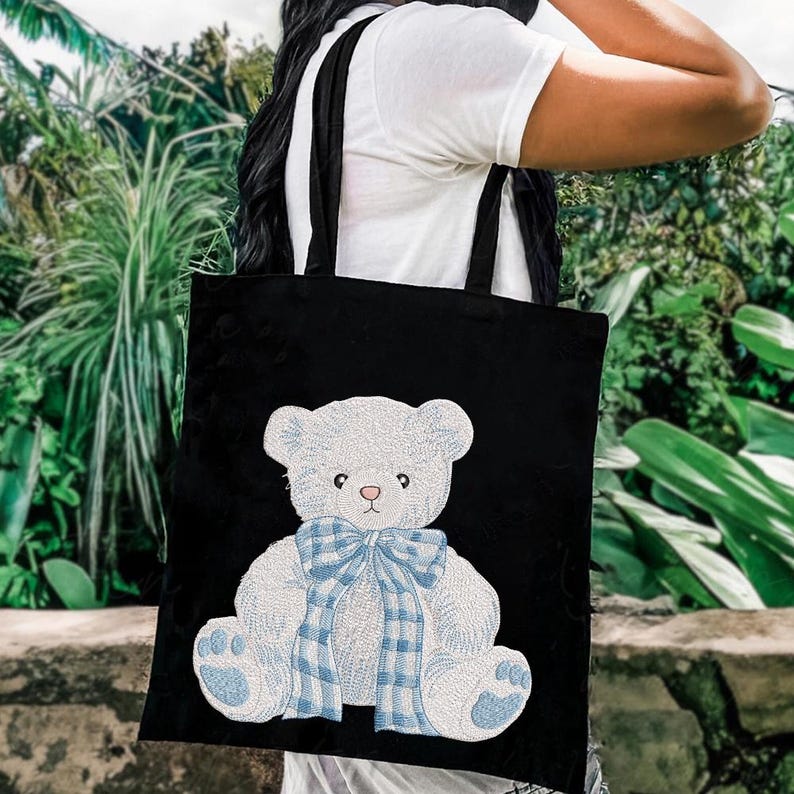 May include: Black tote bag featuring a white embroidered teddy bear with a blue plaid bow and accents. The bag has black straps and is carried over the shoulder. The background is blurred with green foliage.