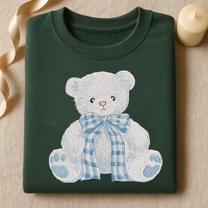 May include: A dark green sweatshirt featuring an embroidered teddy bear design. The bear is white with blue accents, including a plaid bow and paw prints. The sweatshirt has a crew neck and a soft, comfortable appearance.