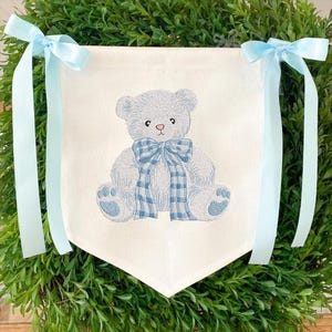 May include: A white banner with a blue embroidered teddy bear wearing a blue plaid bow. Light blue satin ribbons are attached to the top corners. The banner is surrounded by green foliage.