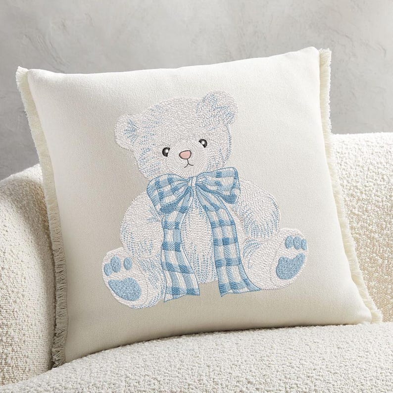 May include: Cream-colored square throw pillow with a detailed embroidered teddy bear design. The bear is white with blue accents, including a plaid bow and paw pads. The pillow has a fringed edge, adding a soft texture to the nursery decor.