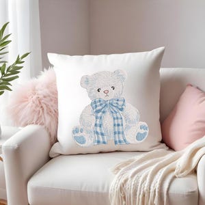 May include: White square pillow with an embroidered teddy bear design. The bear is light blue with a blue plaid bow and accents. The pillow is on a white couch with other decorative pillows.