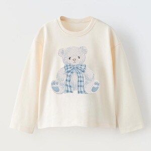 May include: Cream-colored long-sleeve shirt featuring an embroidered teddy bear design. The bear is light blue with a blue and white plaid bow. The shirt has a crew neck and long sleeves, suitable for children's wear.