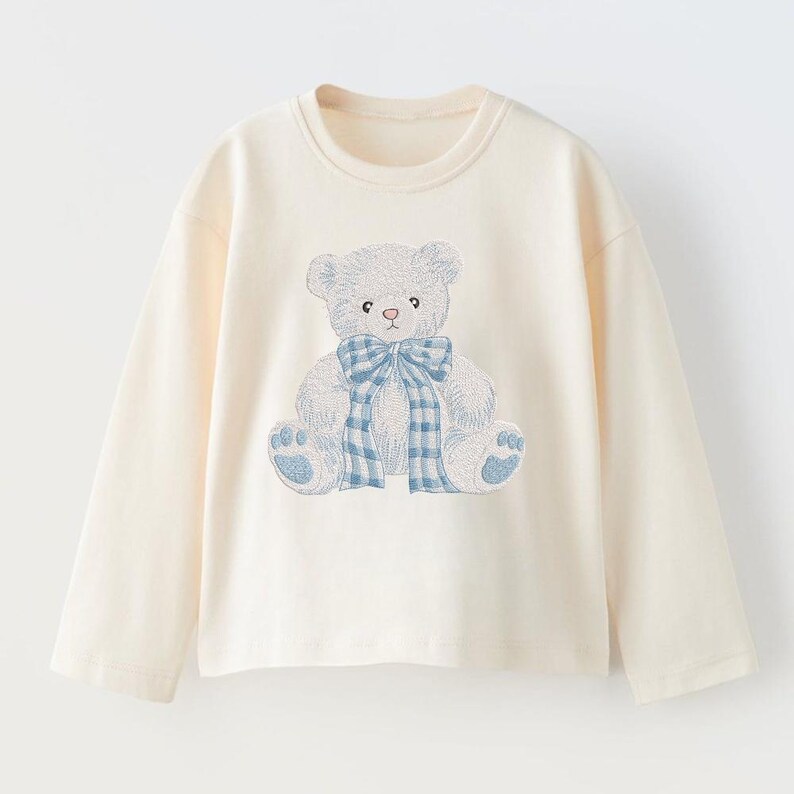May include: Cream-colored long-sleeve shirt featuring an embroidered teddy bear design. The bear is light blue with a blue and white plaid bow. The shirt has a crew neck and long sleeves, suitable for children's wear.