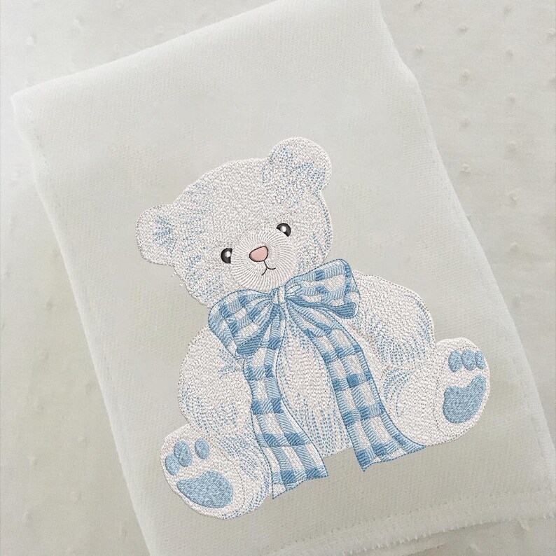 May include: White baby blanket with an embroidered teddy bear design. The teddy bear is light blue with a matching plaid bow and paw prints. The blanket has a textured surface.