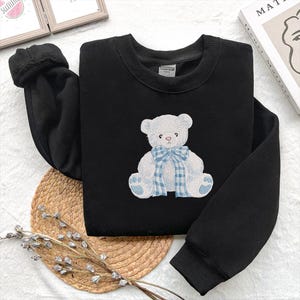 May include: Black sweatshirt featuring a white teddy bear design with a blue plaid bow. The bear is embroidered on the front of the crew neck sweatshirt. The sleeves are long and the material appears soft and comfortable.