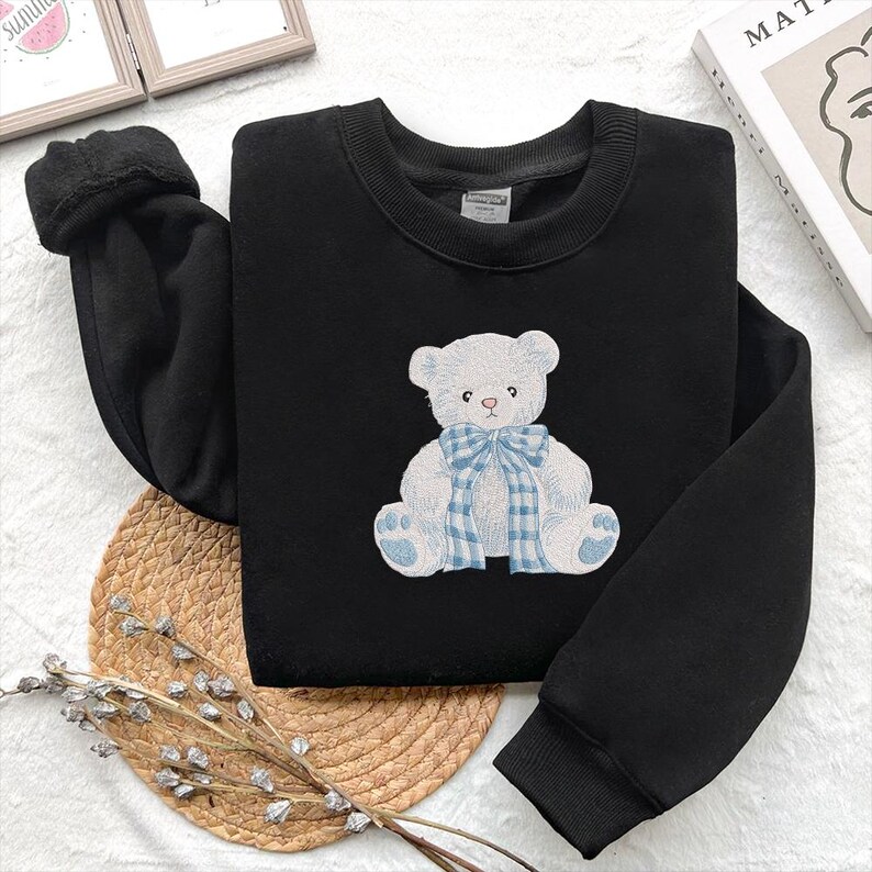 May include: Black sweatshirt featuring a white teddy bear design with a blue plaid bow. The bear is embroidered on the front of the crew neck sweatshirt. The sleeves are long and the material appears soft and comfortable.