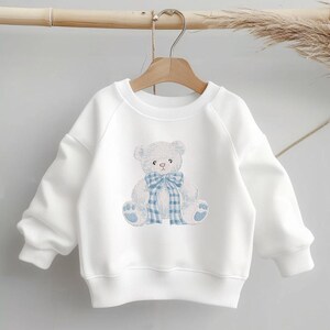 May include: White sweatshirt with a light blue embroidered teddy bear design. The bear has a blue plaid bow and matching paw prints. The sweatshirt is hanging on a wooden hanger.
