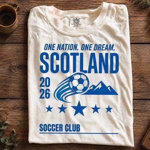 May include: An off-white t-shirt featuring blue text that reads ONE NATION. ONE DREAM. SCOTLAND 20 26 SOCCER CLUB. The graphic includes a soccer ball in motion, a mountain silhouette, and five stars arranged in a horizontal line below the ball.