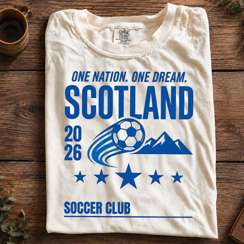 May include: An off-white t-shirt featuring blue text that reads ONE NATION. ONE DREAM. SCOTLAND 20 26 SOCCER CLUB. The graphic includes a soccer ball in motion, a mountain silhouette, and five stars arranged in a horizontal line below the ball.