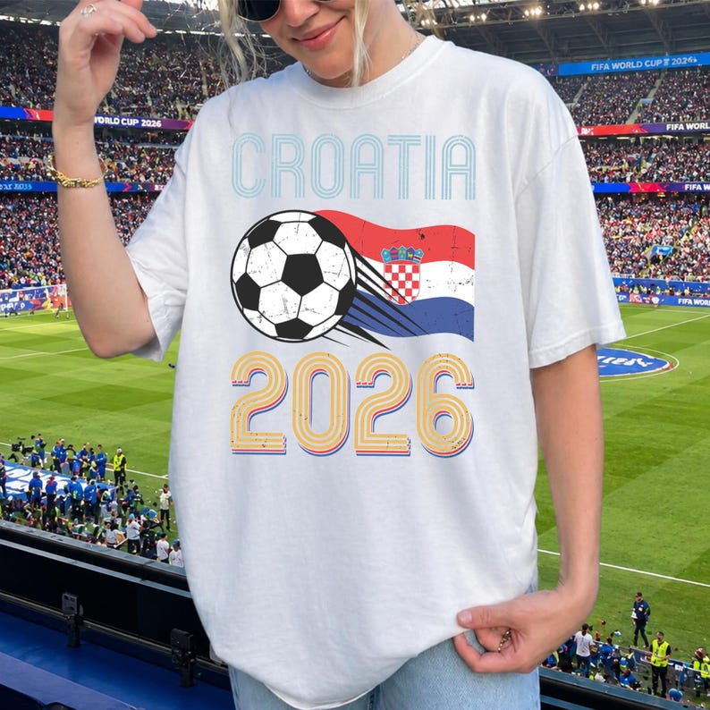 May include: A white t-shirt featuring a graphic print with the text CROATIA in blue, a black and white soccer ball, the Croatian flag, and the year 2026 in large, stylized orange and blue numbers with a distressed, vintage-inspired texture.