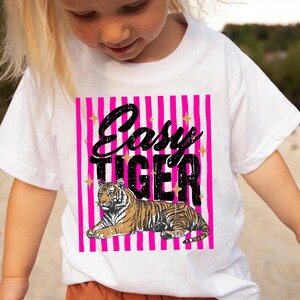 Pu&ograve; includere: T-shirt bianca con un'illustrazione di tigre e la scritta "Easy Tiger". Il design presenta uno sfondo a righe rosa con accenti di stelle dorate. La tigre &egrave; marrone e nera. La t-shirt &egrave; indossata da un bambino.