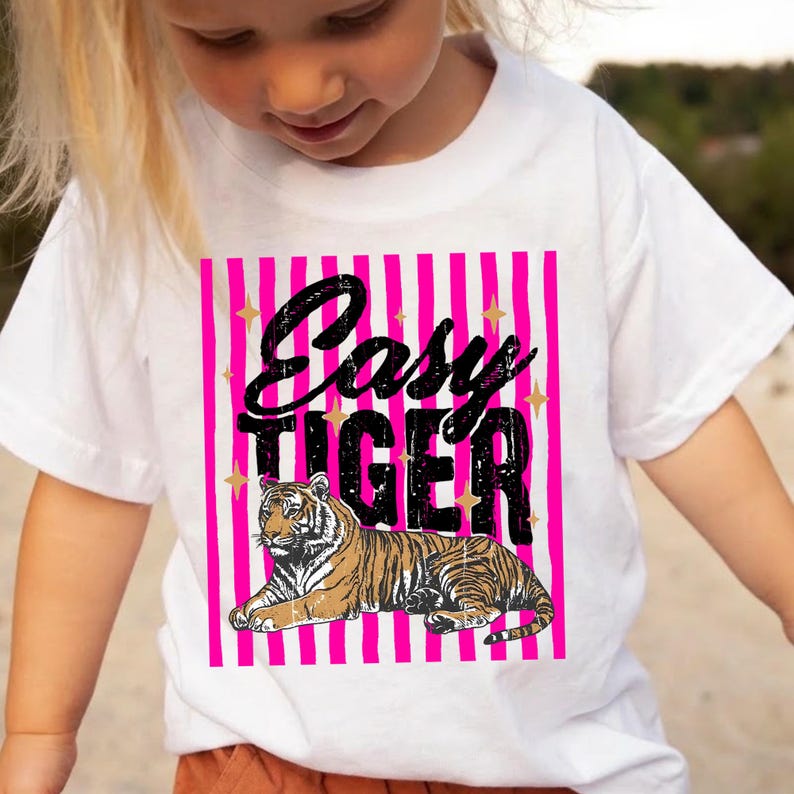 Pu&ograve; includere: T-shirt bianca con un'illustrazione di tigre e la scritta "Easy Tiger". Il design presenta uno sfondo a righe rosa con accenti di stelle dorate. La tigre &egrave; marrone e nera. La t-shirt &egrave; indossata da un bambino.